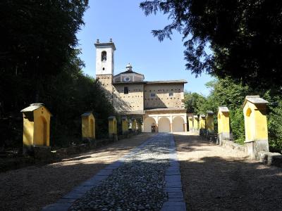 A long, straight processional path lined with yellow chapels leads up to a stone church with a bell tower and a small dome, set against a backdrop of trees and a blue sky.