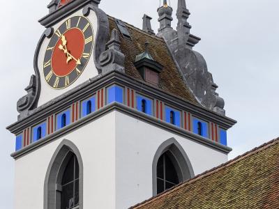 A close-up view of the ornate clock tower of the Stadtkirche Aarau, featuring a red clock face with gold Roman numerals, blue and red decorative panels, and a tiled roof against a cloudy sky.