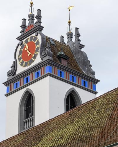A close-up view of the ornate clock tower of the Stadtkirche Aarau, featuring a red clock face with gold Roman numerals, blue and red decorative panels, and a tiled roof against a cloudy sky.