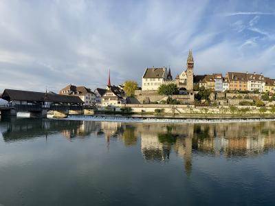 A panoramic view of a historic Swiss town on a riverbank featuring a prominent covered wooden bridge, a stone tower, and several church spires reflected in the water.
