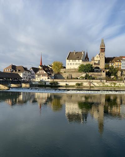 A panoramic view of a historic Swiss town on a riverbank featuring a prominent covered wooden bridge, a stone tower, and several church spires reflected in the water.