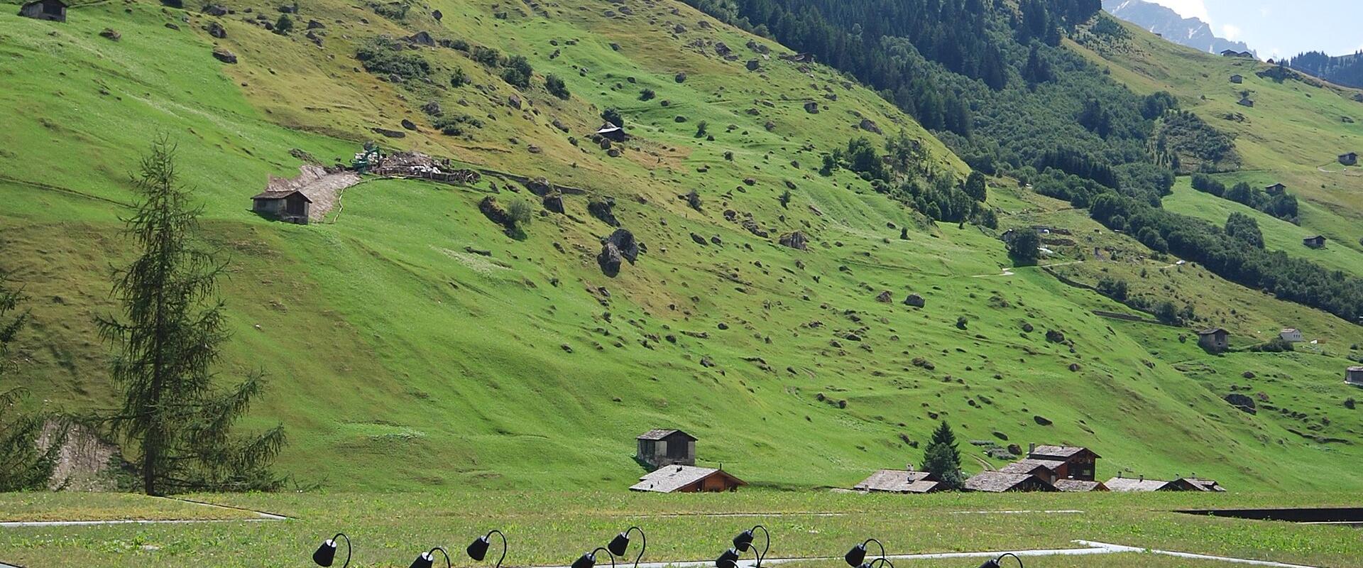 A grassy outdoor area with a grid of black, curved light fixtures mounted over rectangular glass panels set into the ground, with a steep green alpine slope and scattered chalets in the background.