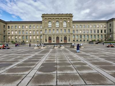 A wide-angle view of the ETH Zurich Main Building (Hauptgebäude) facing the Polyterrasse plaza, with its distinctive neoclassical facade, central tower, and symmetrical wings; people are scattered across the paved square, some sitting on benches o