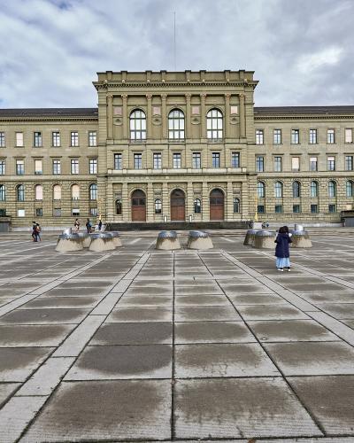 A wide-angle view of the ETH Zurich Main Building (Hauptgebäude) facing the Polyterrasse plaza, with its distinctive neoclassical facade, central tower, and symmetrical wings; people are scattered across the paved square, some sitting on benches o