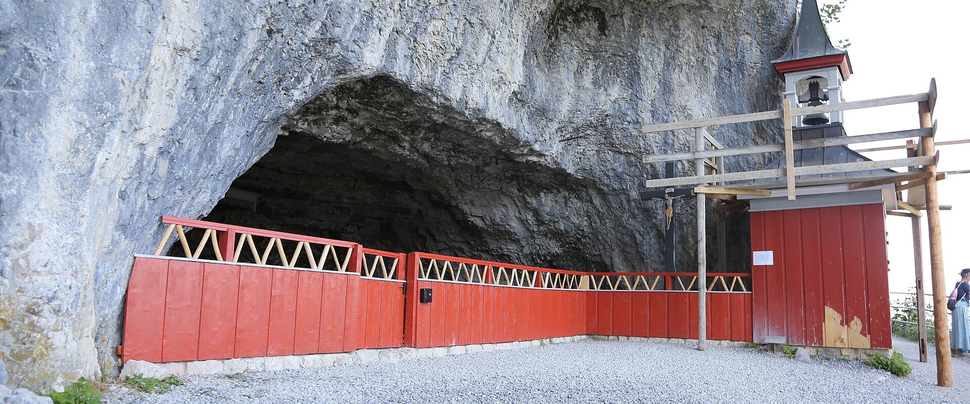 The image shows the entrance to a large limestone cave featuring a distinctive red wooden fence and a small wooden chapel with a bell tower built directly into the rock face.