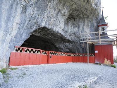 The image shows the entrance to a large limestone cave featuring a distinctive red wooden fence and a small wooden chapel with a bell tower built directly into the rock face.