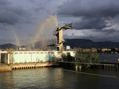 A concrete pier structure with a tall diving tower, a low building with light blue panels, and a rainbow over the water with mountains in the background.