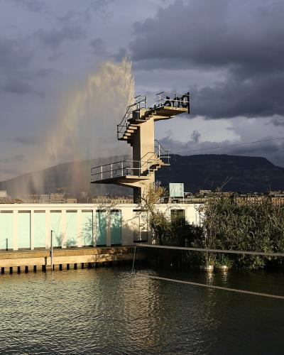 A concrete pier structure with a tall diving tower, a low building with light blue panels, and a rainbow over the water with mountains in the background.