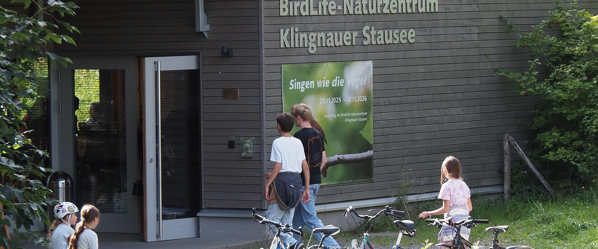 The image shows the exterior of a modern wooden building with a green roof, featuring large signage that reads "BirdLife-Naturzentrum Klingnauer Stausee" above an entrance where several people and bicycles are gathered.