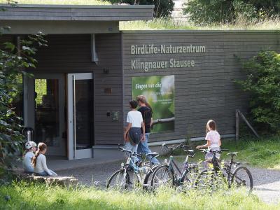 The image shows the exterior of a modern wooden building with a green roof, featuring large signage that reads "BirdLife-Naturzentrum Klingnauer Stausee" above an entrance where several people and bicycles are gathered.