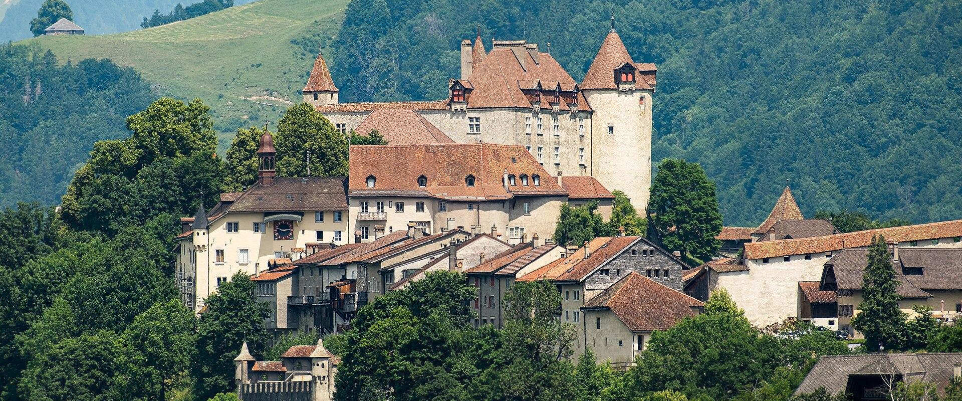 A large medieval castle with distinctive conical towers and terracotta roofs sits atop a hill overlooking a cluster of traditional buildings and a fortified wall, set against a backdrop of green forested mountains.