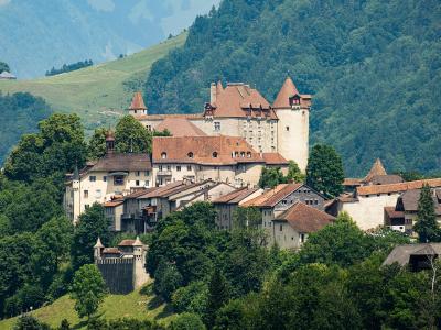 A large medieval castle with distinctive conical towers and terracotta roofs sits atop a hill overlooking a cluster of traditional buildings and a fortified wall, set against a backdrop of green forested mountains.