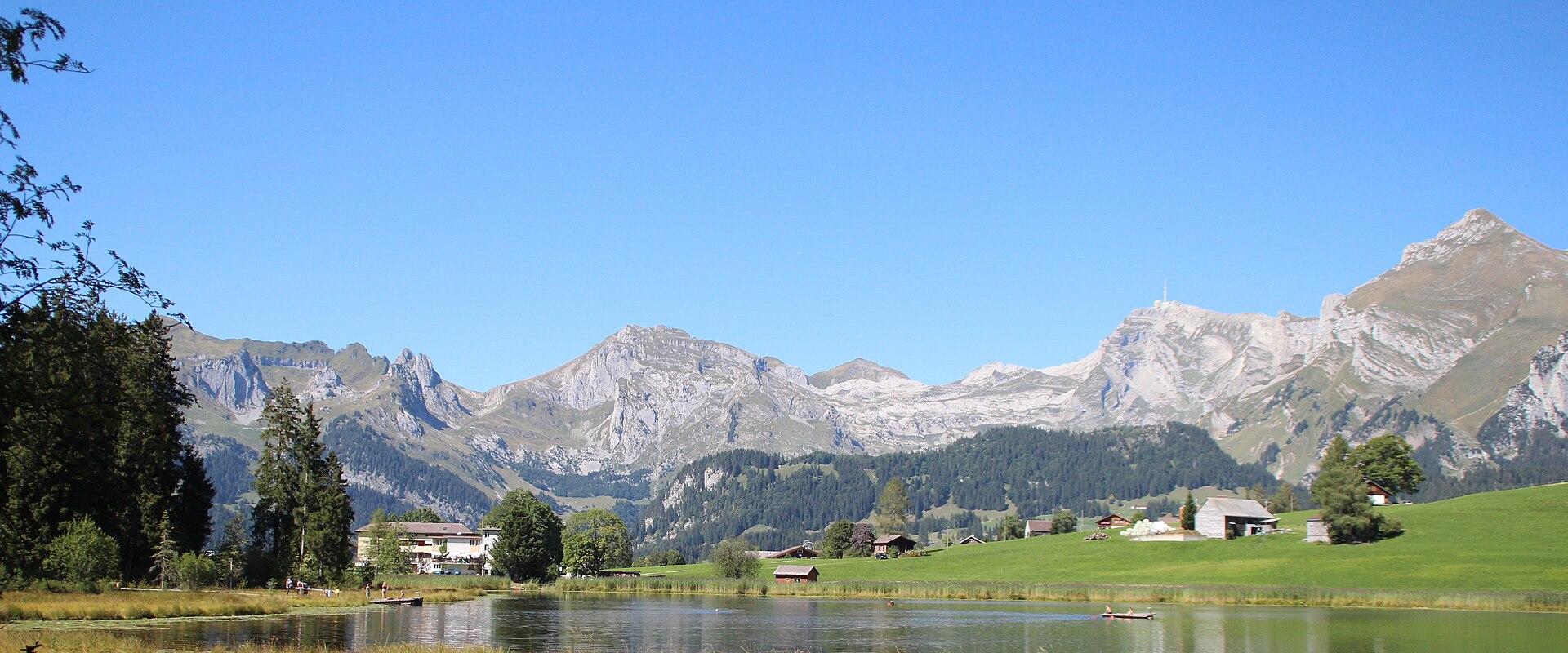 A landscape photograph showing a calm lake in the foreground with green pastures, scattered farm buildings, and a prominent mountain range with a distinct peak featuring a communications tower on the right side under a clear blue sky.