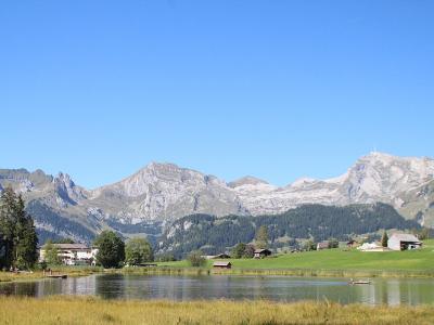 A landscape photograph showing a calm lake in the foreground with green pastures, scattered farm buildings, and a prominent mountain range with a distinct peak featuring a communications tower on the right side under a clear blue sky.