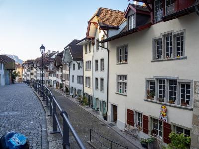 A cobblestone street lined with traditional multi-story residential buildings featuring white facades, red shutters, and tiled roofs, viewed from a slightly elevated perspective with a metal railing in the foreground.