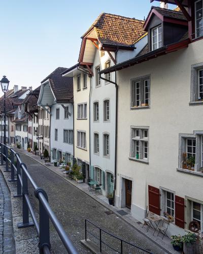 A cobblestone street lined with traditional multi-story residential buildings featuring white facades, red shutters, and tiled roofs, viewed from a slightly elevated perspective with a metal railing in the foreground.