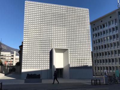 A tall, modern building with a distinctive white grid-patterned facade and a recessed entrance, situated in an urban plaza with mountains in the background.