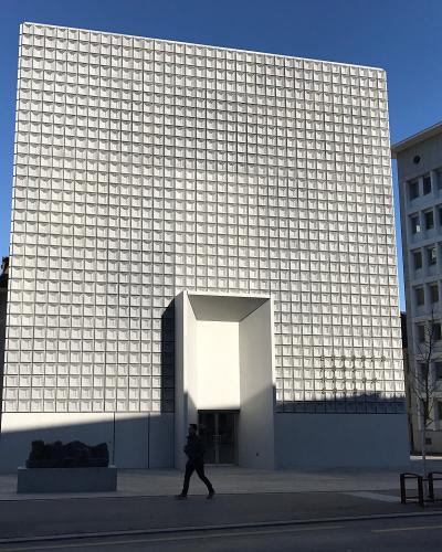 A tall, modern building with a distinctive white grid-patterned facade and a recessed entrance, situated in an urban plaza with mountains in the background.