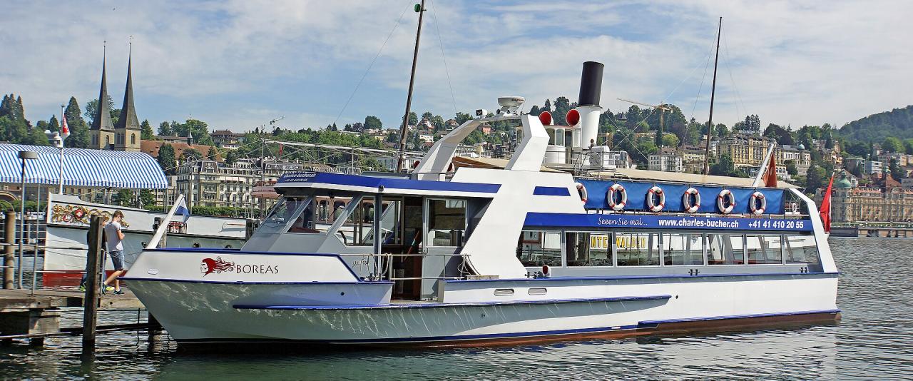 A white and blue paddle steamer-style boat named 'BOREAS' is docked at a pier, with a town featuring twin-spired churches and hillside buildings in the background.