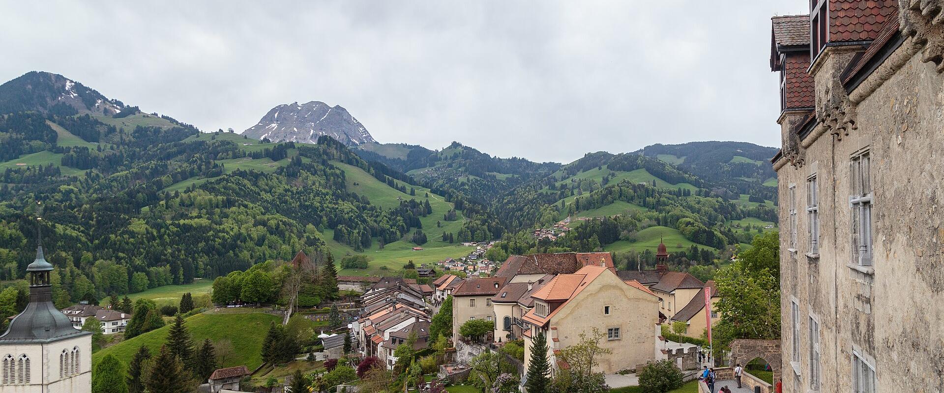 A view from a stone castle wall looking down over the rooftops of a historic town, with green hills and mountains in the background and a glider in the sky.