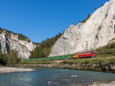 A red and orange Rhaetian Railway freight train hauling green "VALSER" wagons along a track beside a river, with steep limestone cliffs rising on either side.
