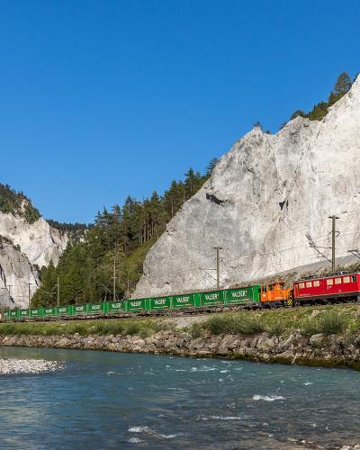 A red and orange Rhaetian Railway freight train hauling green "VALSER" wagons along a track beside a river, with steep limestone cliffs rising on either side.