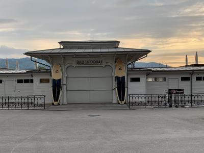 A white wooden building with a sign reading "BAD UTOQUAI" and two surfboards mounted on either side of the entrance, set against a backdrop of hills and a cloudy sky.