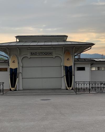 A white wooden building with a sign reading "BAD UTOQUAI" and two surfboards mounted on either side of the entrance, set against a backdrop of hills and a cloudy sky.