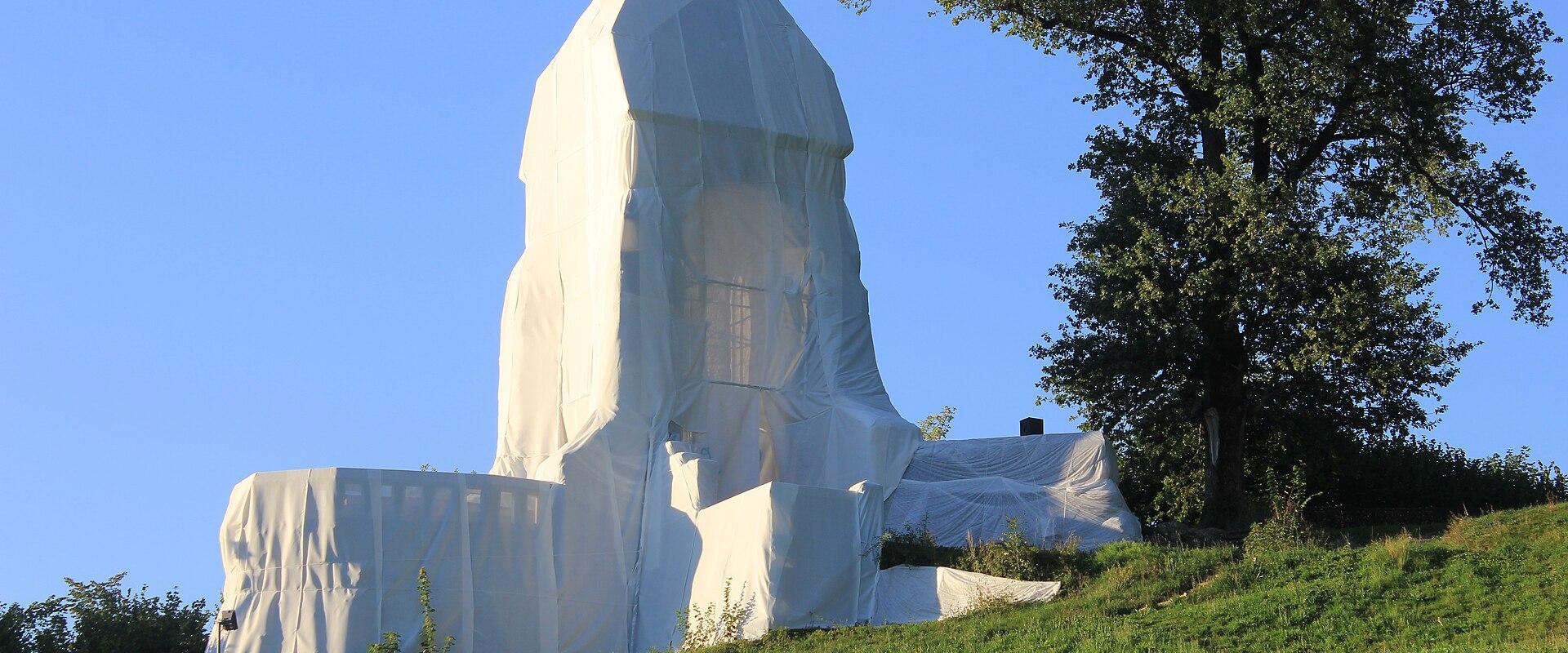 A large stone monument covered in white protective sheeting and scaffolding, situated on a grassy hillside next to a tree.