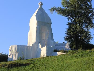 A large stone monument covered in white protective sheeting and scaffolding, situated on a grassy hillside next to a tree.