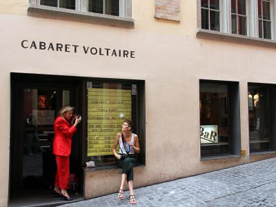 The image shows the exterior of a building with "CABARET VOLTAIRE" in large black letters above the entrance, two people standing near the doorway, and a yellow poster listing events inside the window; the street is paved with cobblestones and the