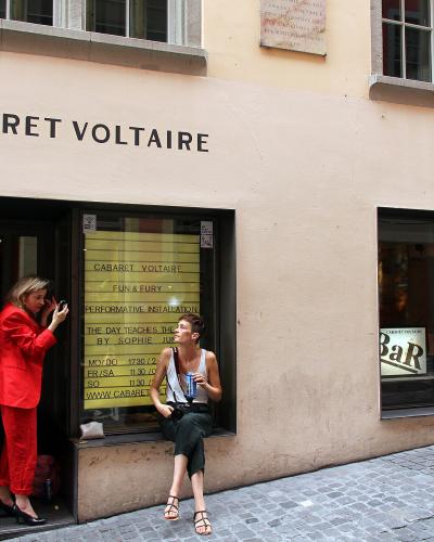 The image shows the exterior of a building with "CABARET VOLTAIRE" in large black letters above the entrance, two people standing near the doorway, and a yellow poster listing events inside the window; the street is paved with cobblestones and the