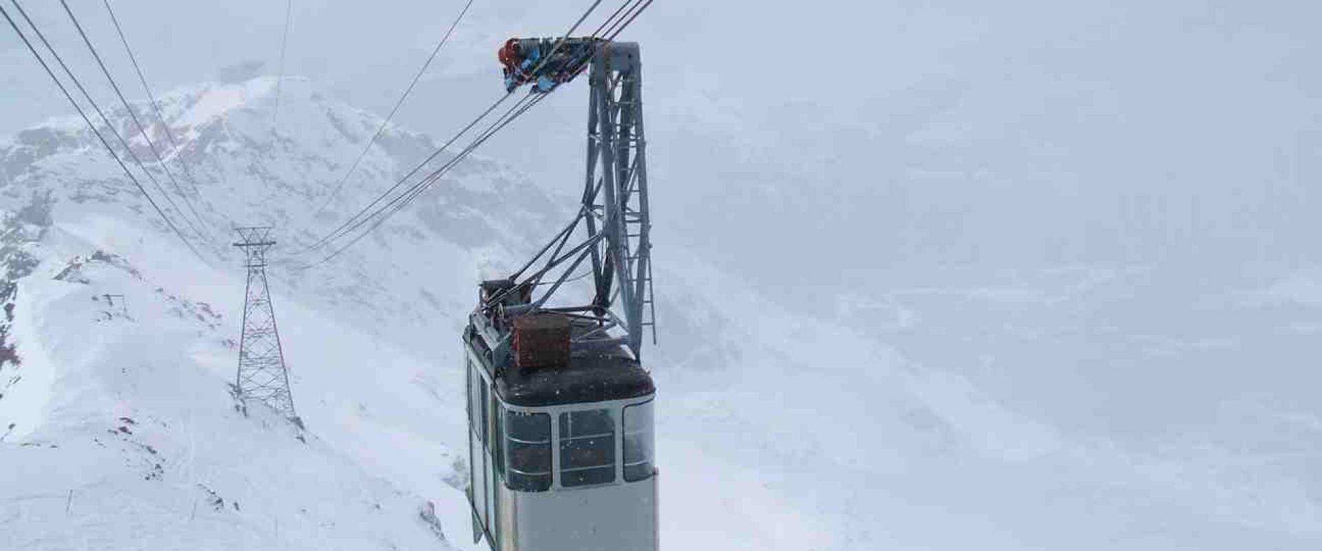 A cable car gondola suspended on cables against a snowy mountain backdrop, with a worker visible on the overhead mechanism.
