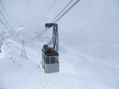 A cable car gondola suspended on cables against a snowy mountain backdrop, with a worker visible on the overhead mechanism.