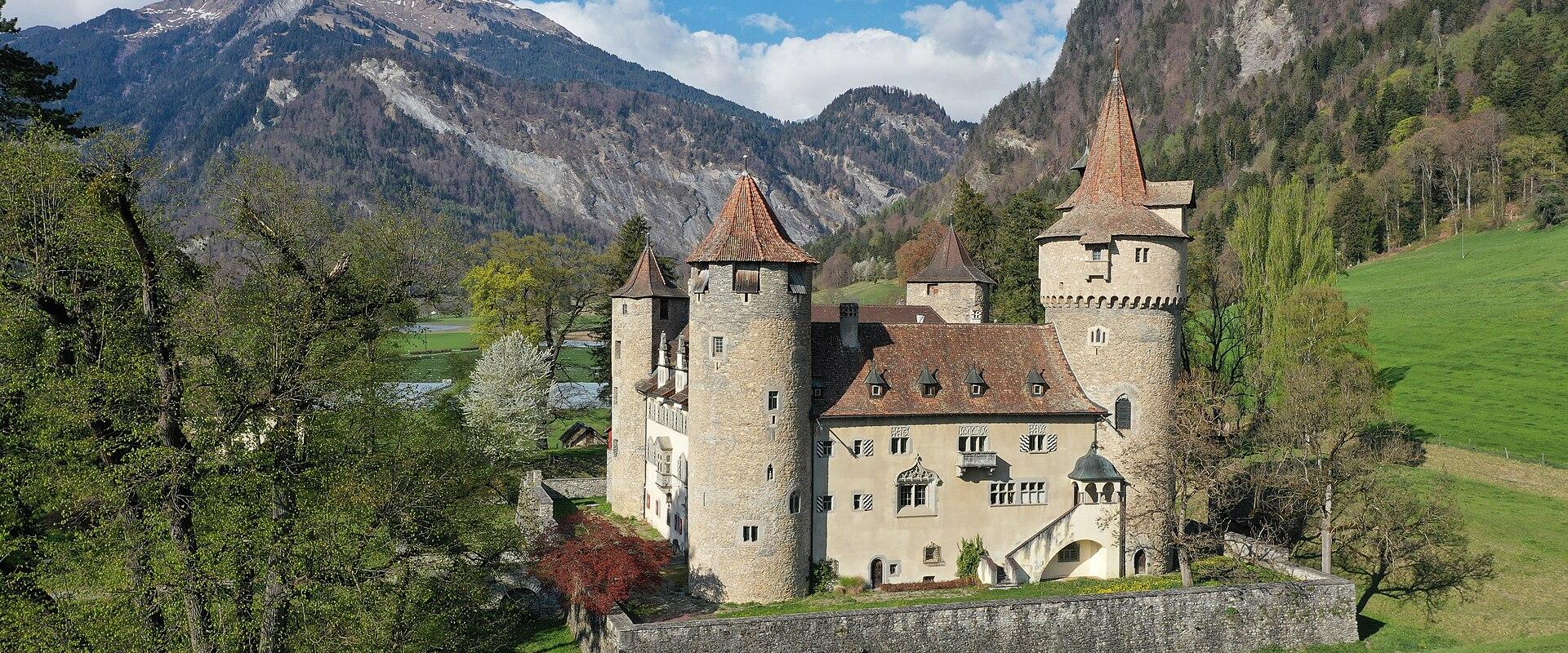 A medium shot of a historic castle with multiple towers and a terracotta-tiled roof, situated in a green, mountainous landscape with a cow grazing in the foreground.