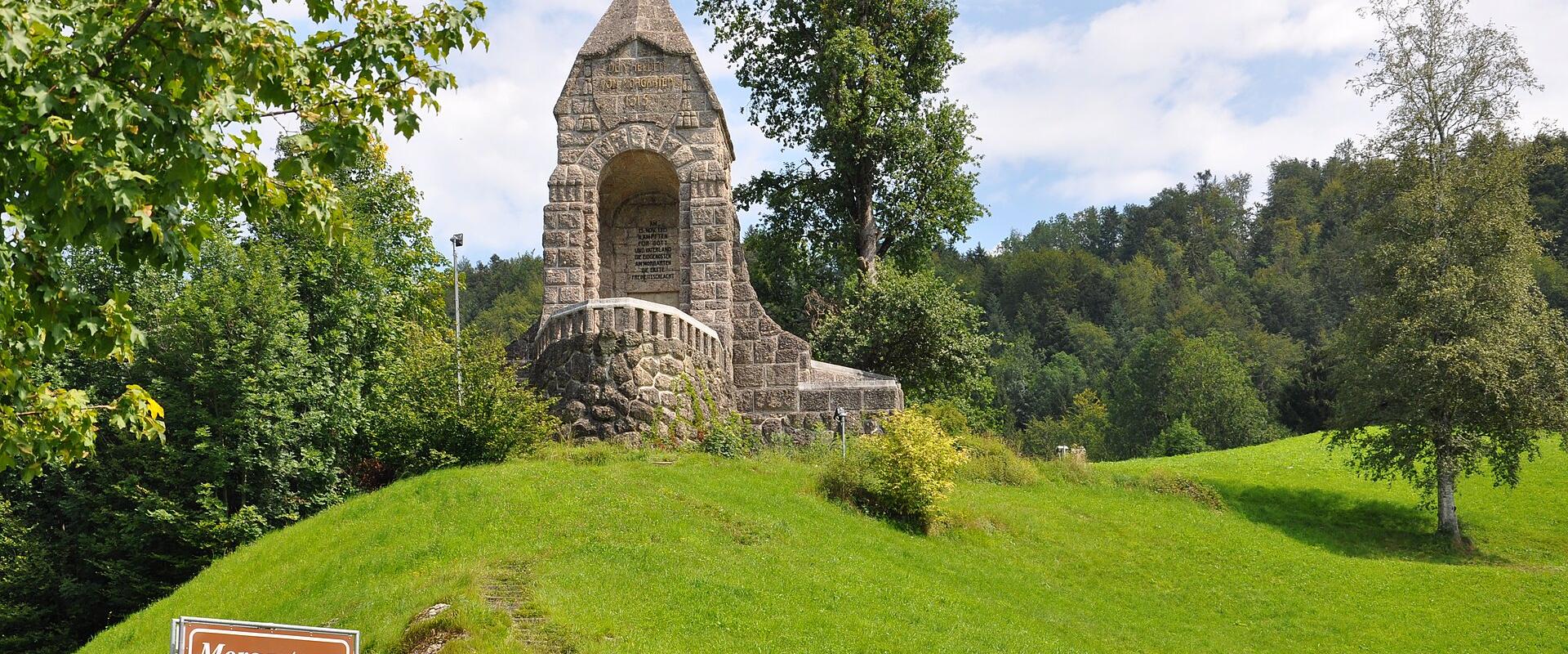 A large stone monument on a grassy hillside, currently covered in white protective sheeting and scaffolding, with a cross visible at the top.