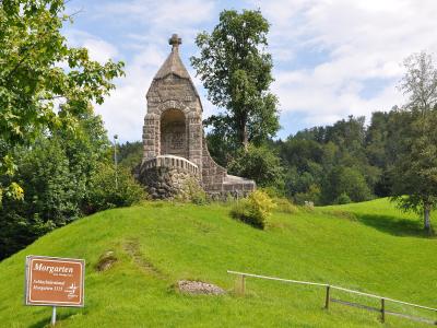 A large stone monument on a grassy hillside, currently covered in white protective sheeting and scaffolding, with a cross visible at the top.
