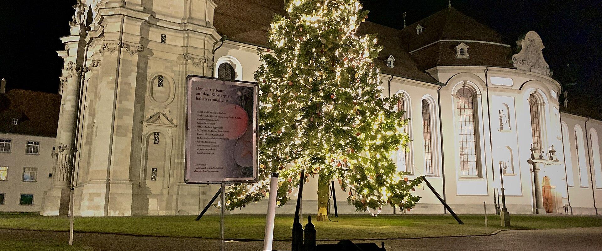 A night view of a large white Baroque church with a prominent clock tower, featuring a lit Christmas tree and a sign in the foreground.