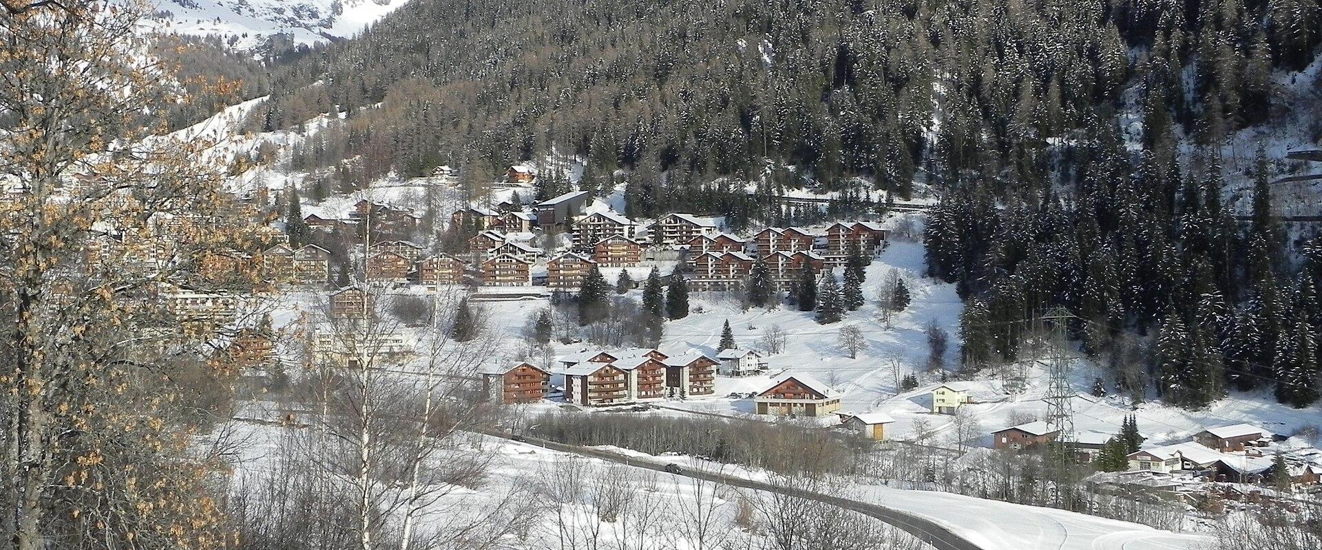 A snowy mountain valley scene showing a cluster of chalet-style buildings and a road winding through the snow, with forested slopes and peaks in the background.