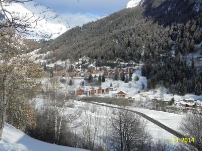A snowy mountain valley scene showing a cluster of chalet-style buildings and a road winding through the snow, with forested slopes and peaks in the background.