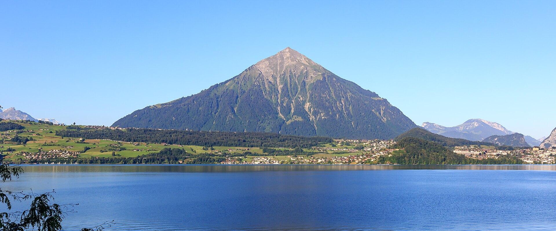A wide landscape shot showing a large, pyramid-shaped mountain rising above a lake and a town on the shore under a clear blue sky.