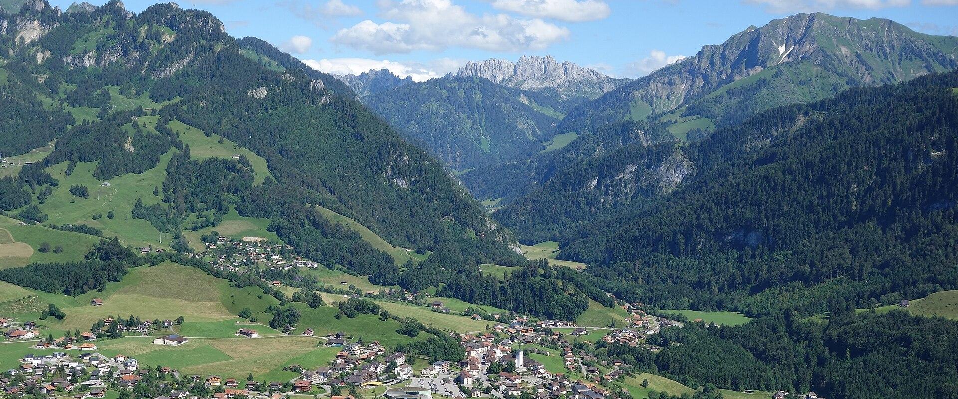 A high-angle, wide landscape photograph showing a village nestled in a green valley, surrounded by steep, forested mountains, with a prominent rocky peak (the Dapples) visible in the background.