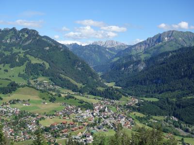 A high-angle, wide landscape photograph showing a village nestled in a green valley, surrounded by steep, forested mountains, with a prominent rocky peak (the Dapples) visible in the background.