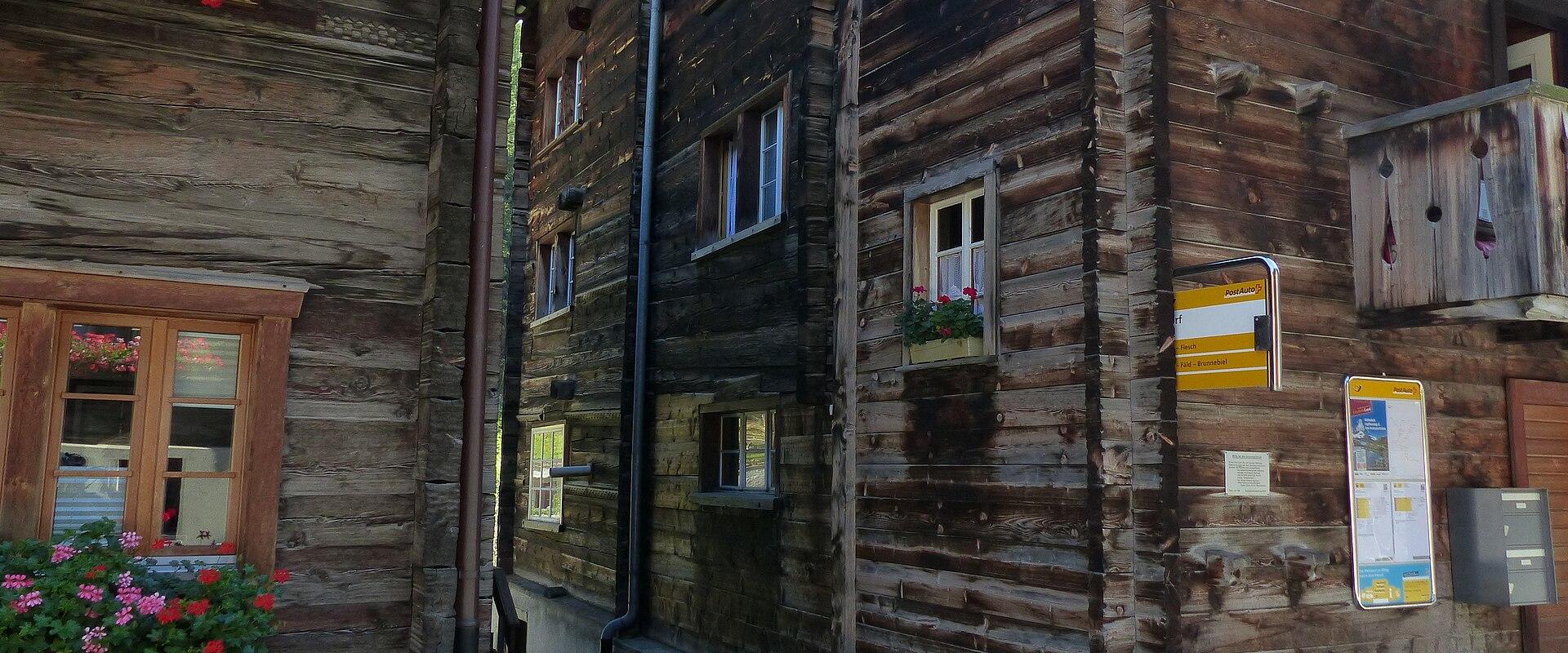 A close-up view of traditional wooden chalet-style buildings in a narrow village street, featuring flower boxes with pink and red geraniums, a yellow information sign, and a notice board mounted on the wall.