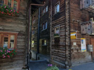A close-up view of traditional wooden chalet-style buildings in a narrow village street, featuring flower boxes with pink and red geraniums, a yellow information sign, and a notice board mounted on the wall.