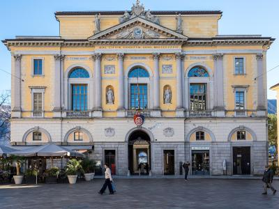 A large, yellow neoclassical building with a central pediment, statues, and the Latin inscription "AERE CIVIVM CONDITVM ANNO MDCCCXXXIV" above the entrance, situated in a paved public square with people walking and outdoor cafe seating.