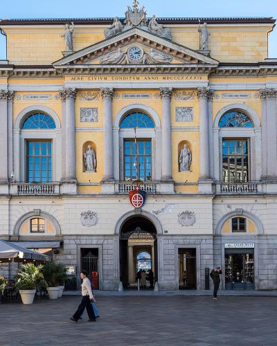 A large, yellow neoclassical building with a central pediment, statues, and the Latin inscription "AERE CIVIVM CONDITVM ANNO MDCCCXXXIV" above the entrance, situated in a paved public square with people walking and outdoor cafe seating.