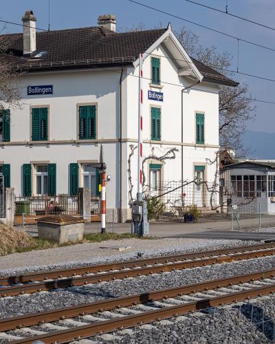 A white two-story railway station building with green shutters, situated next to train tracks, featuring blue signs that read "Bollingen" on the facade and a vertical pole.