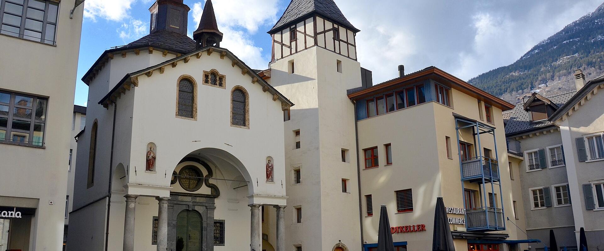 A white chapel with a central arched entrance, two small spires, and a tall adjacent tower stands in a paved town square next to a building labeled "RESTAURANT COLUMBUS" and "DINKELLER", with mountains in the background.