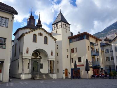 A white chapel with a central arched entrance, two small spires, and a tall adjacent tower stands in a paved town square next to a building labeled "RESTAURANT COLUMBUS" and "DINKELLER", with mountains in the background.